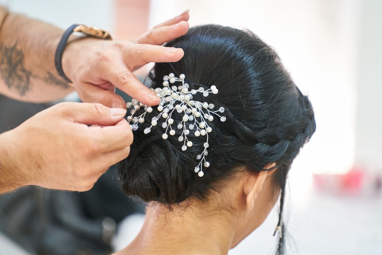 hero-img-01 Photograph of a hairstylist arranging an elegant updo embellished with delicate decorative pins in a professional studio.