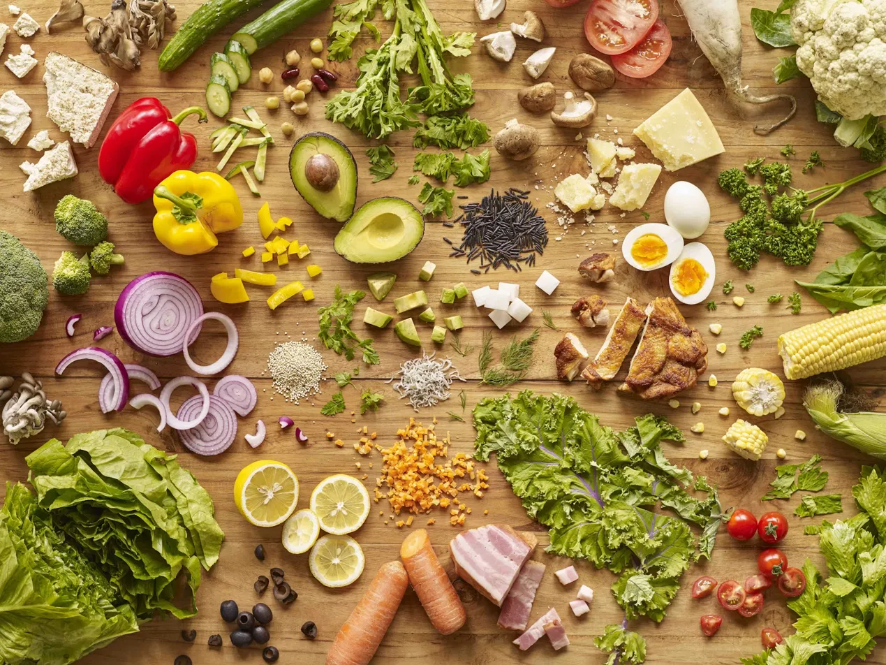 Overhead flat lay of fresh healthy foods on wooden surface: broccoli, leafy greens, avocado, red and yellow peppers, eggs, mushrooms, tomatoes, cucumber, carrots, onion, nuts, cheese, whole grains, lemon, and herbs