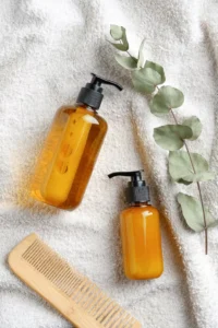 Overhead view of essential hair care products arranged on a white towel: amber-colored shampoo and conditioner bottles with pump dispensers, a wooden wide-tooth comb, and eucalyptus leaves, representing budget-friendly products for maintaining braided ponytail hairstyles